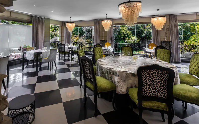 Elegant dining room with green velvet chairs, round draped table, crystal chandeliers, black-and-white checkered floor, and garden views.