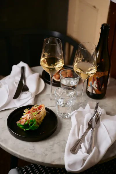 Tabletop dining setup with green salad on black plate, white wine glasses, beer bottle, and utensils on white napkins.
