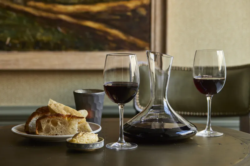 Tabletop with crusty bread, butter, red wine in glasses and decanter, painting and green chair in cozy inn background