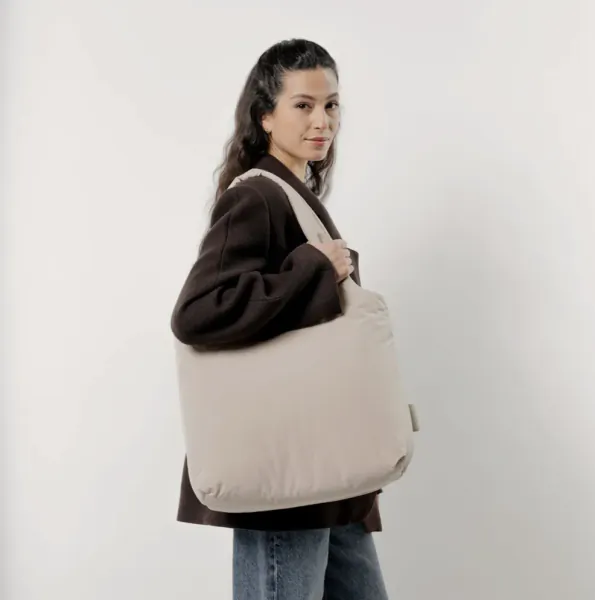 Woman with wavy hair smiling, holding neutral sandstone Kapten & Son tote bag, wearing brown cardigan and jeans