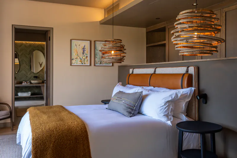 Cozy hotel bedroom with tan leather headboard, white bed, orange throw, rattan pendant lights, and en-suite bathroom in Addo.