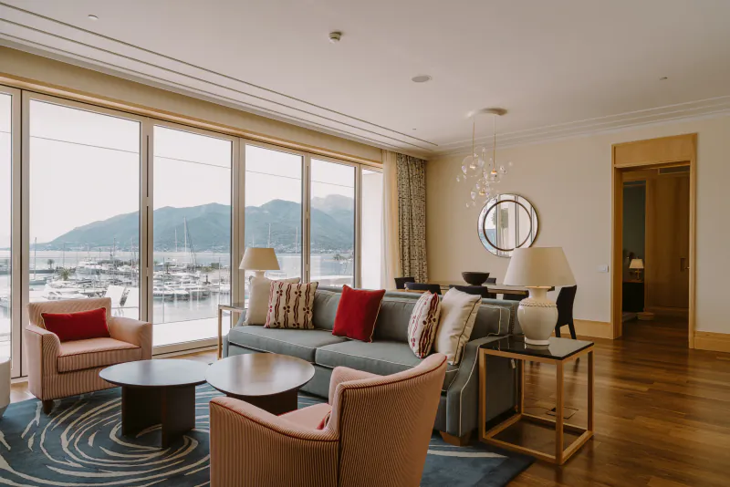 Luxurious hotel living room with gray sofa, red armchairs, coffee tables, and panoramic view of marina and mountains.