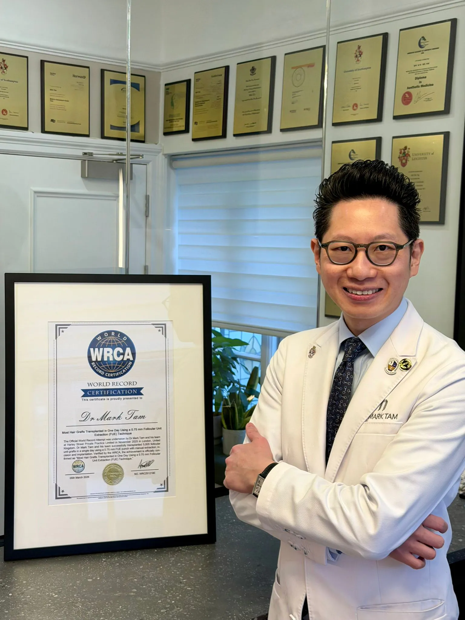 Man in white suit and glasses smiles confidently beside framed WRCA 'Dr. Min Tan' award, with trophy wall behind.