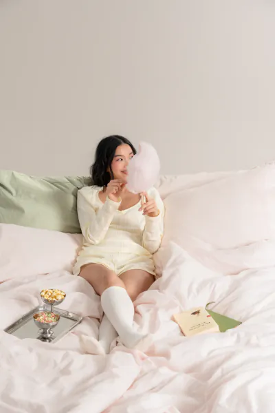 Woman in soft yellow knit set lounging on bed, holding pink cotton candy, with yogurt tray nearby