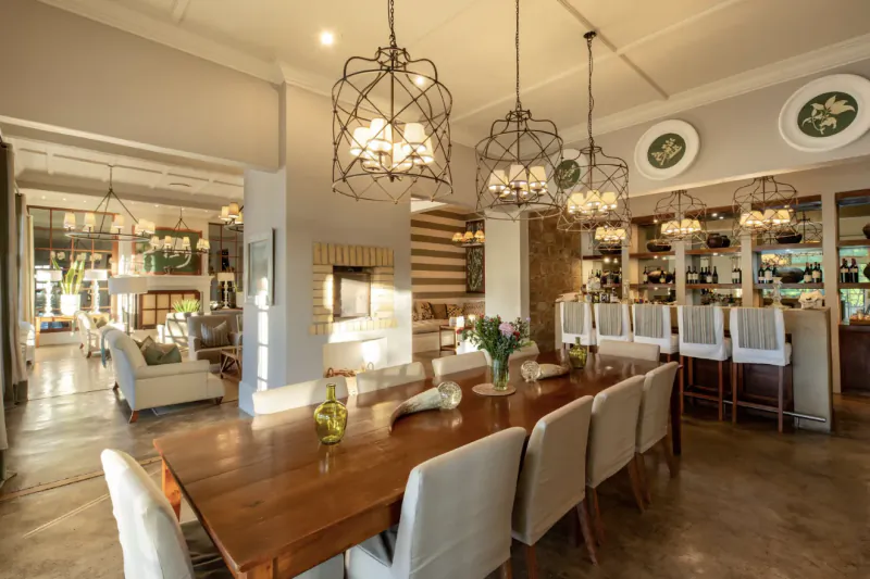 Elegant dining room in Samara Karoo Safari Villa with long wooden table, white chairs, bar, pendant lights, and fireplace