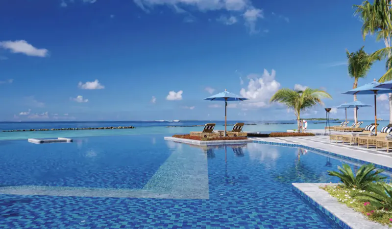 Infinity pool overlooking turquoise ocean, blue umbrellas, lounge chairs, palm trees, clear blue sky