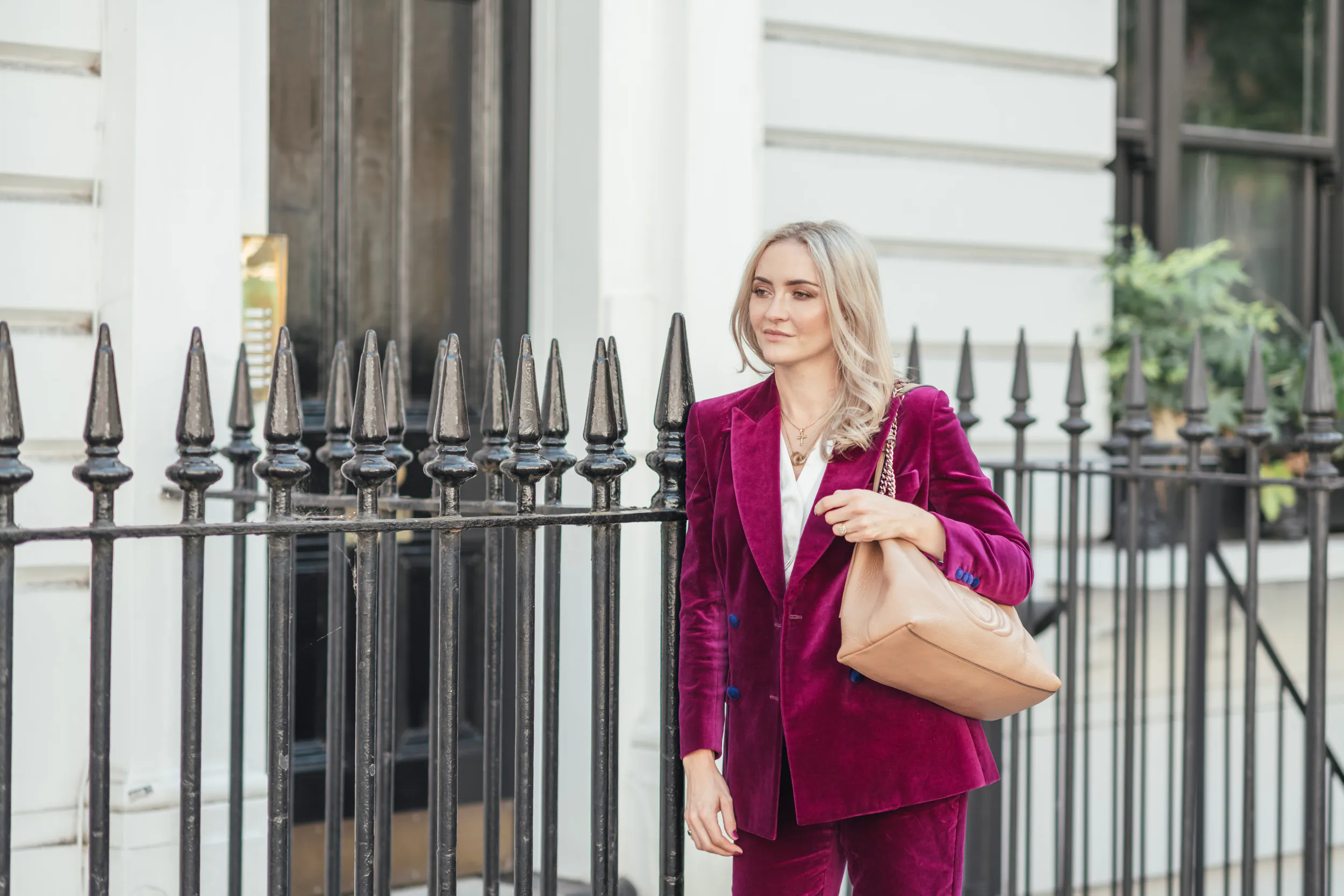 Dr Catharine Denning in pink velvet suit and beige bag, walking by ornate black iron fence outside white building