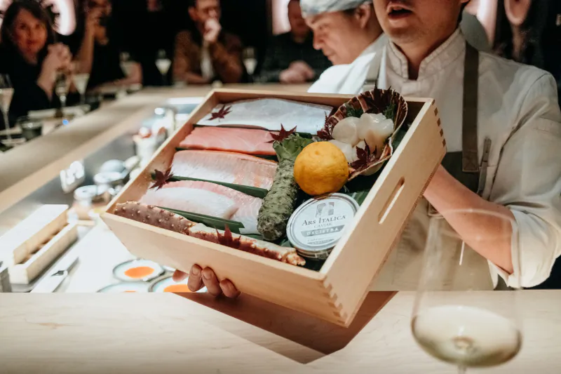 Chef holding wooden box of fresh sashimi slices, garnished with lemon and herbs, at LUNA Omakase sushi bar