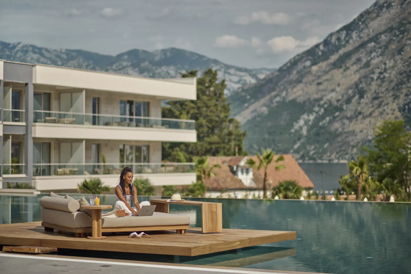 Woman in white sits on infinity pool deck lounger at Hyatt Regency Kotor Bay Resort, Montenegro mountains and bay backdrop.