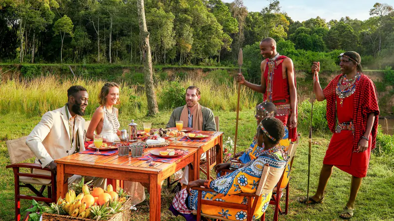 Diverse group dining at wooden table with Kenyan Maasai men in red attire, surrounded by lush riverside greenery and fruit bowl.