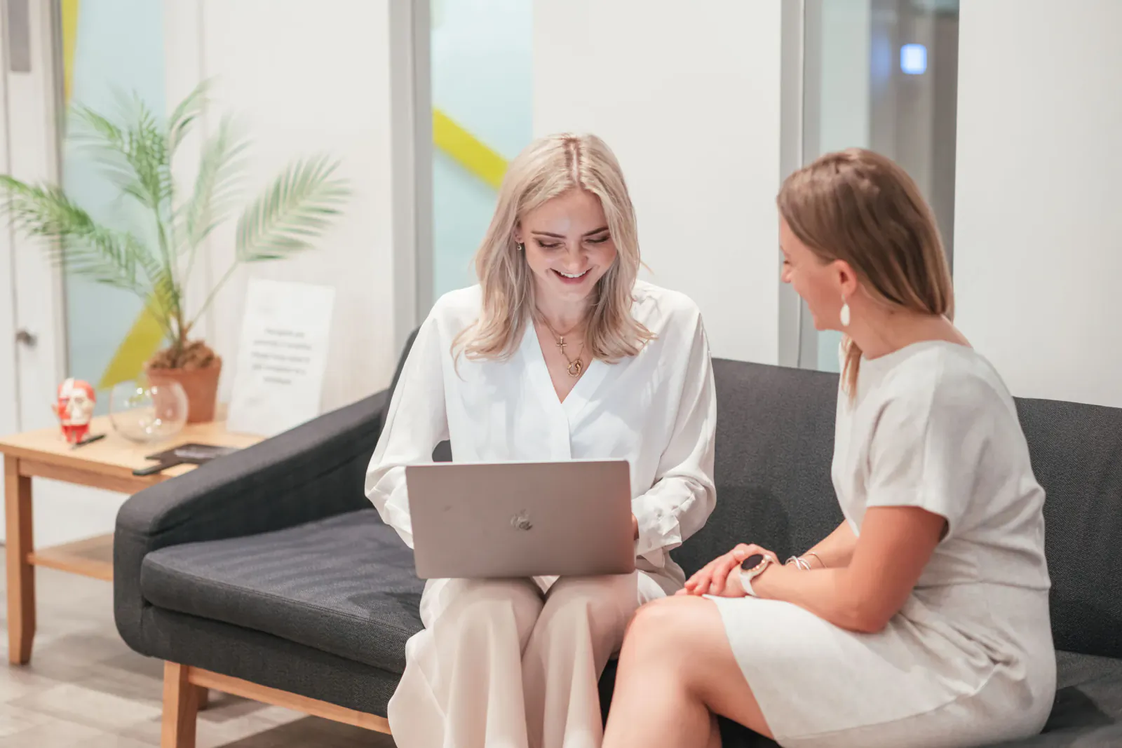 Two smiling women in office attire seated on black sofa, one using laptop, plants and side table nearby