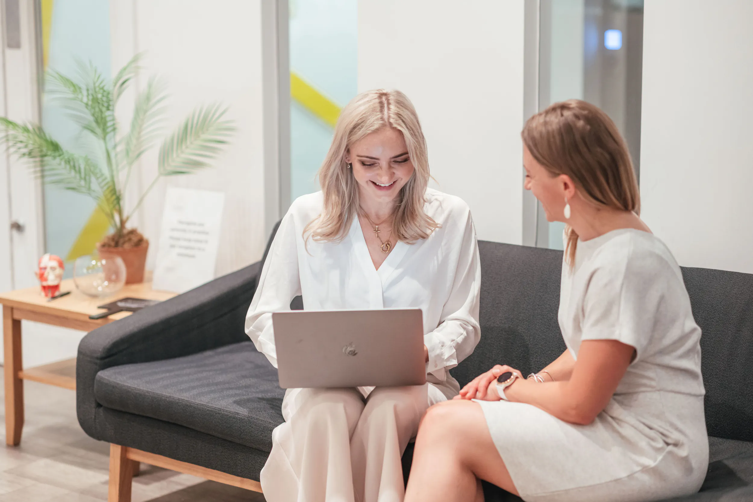 Two smiling women in office attire seated on black sofa, one using laptop, plants and side table nearby