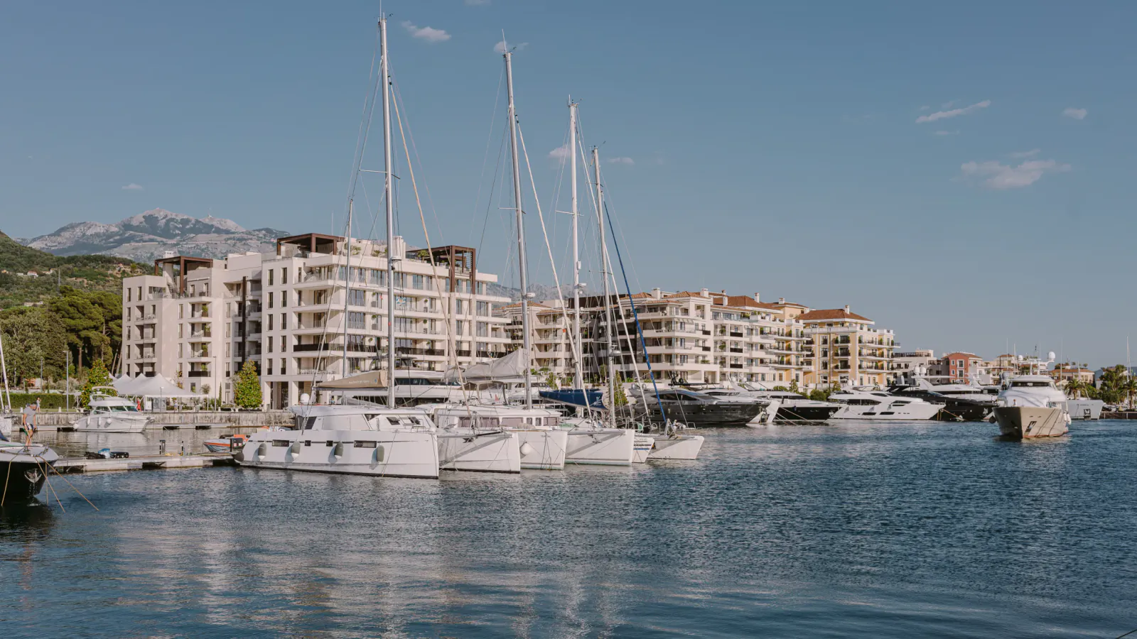 Luxury yachts docked in Porto Montenegro marina, modern white buildings, clear blue water, distant mountains under blue sky.