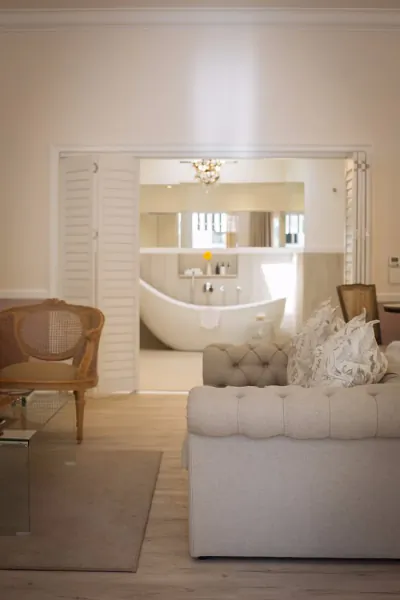 Elegant living room with white tufted sofa and rattan chair opening to luxurious bathroom with freestanding bathtub.