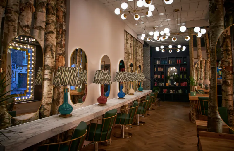 Edward James London hair salon interior with birch tree walls, green stylist chairs, mirrors, lamps, and globes.