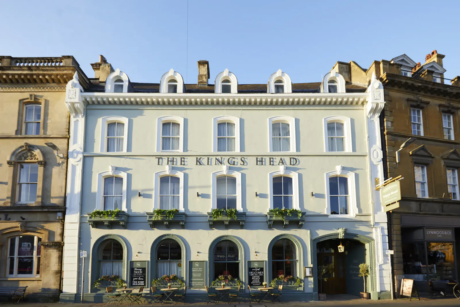 The Kings Head pub, a charming pale green Georgian building with flower boxes on a sunny street in Cirencester, Cotswolds.