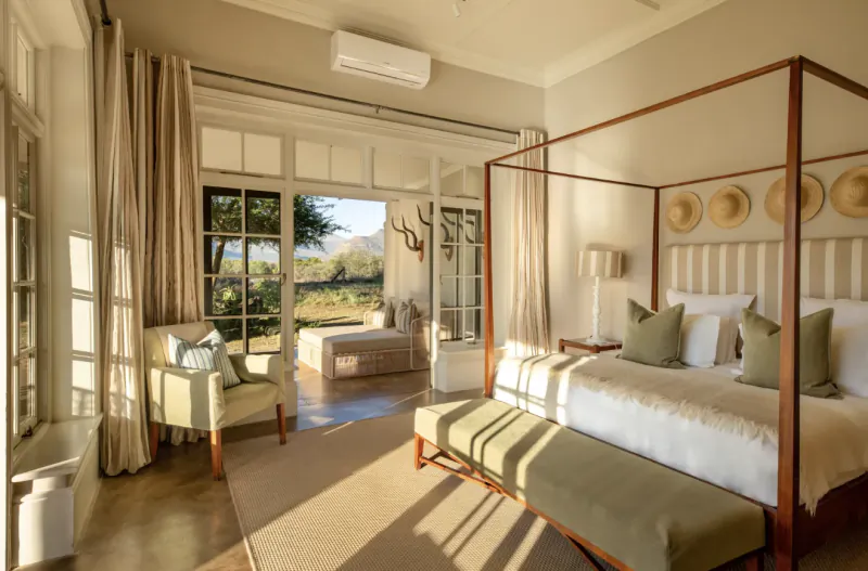 Elegant bedroom in Samara Karoo Safari Villa with four-poster bed, garden view, and straw hats on wall.