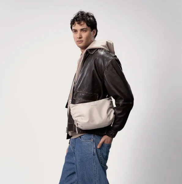 Man in black leather jacket, white hoodie, blue jeans, and neutral sandstone crossbody bag, side pose on white background