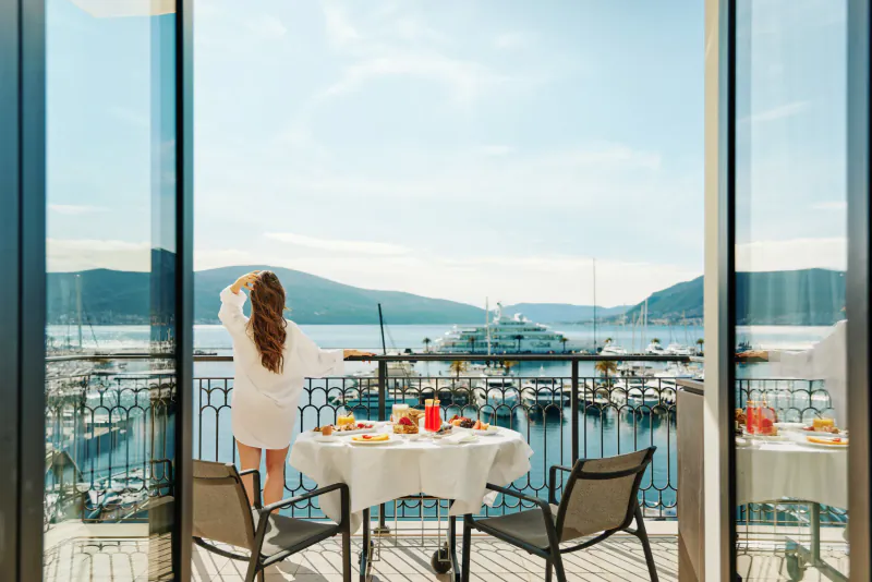 Woman in white robe stands on balcony overlooking marina with yacht, mountains, sea, and breakfast table