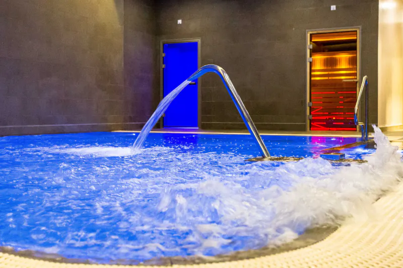 Blue-lit indoor pool with curved silver fountain jet spraying water, blue door and sauna nearby.