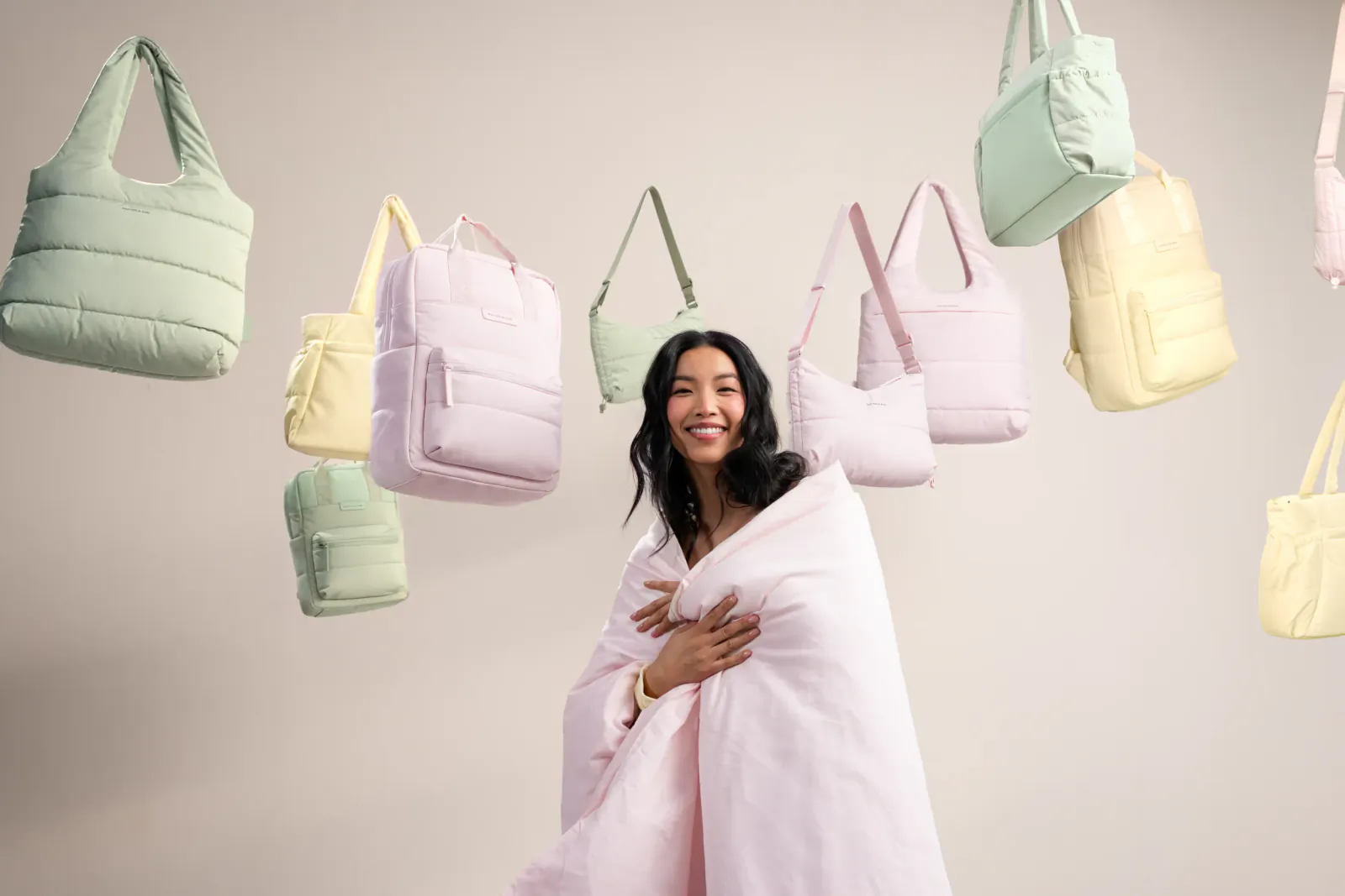 Smiling Asian woman wrapped in pink blanket, surrounded by floating puffy pastel green, pink, yellow bags from Kapten & Son Cloud Club.