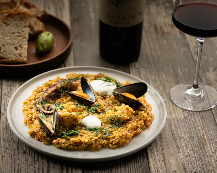 Plate of yellow risotto with octopus, mussels, and dill, beside bread, Acantis wine bottle, and red wine glass on wooden table.
