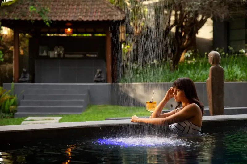 Woman with long dark hair sits pensively in blue-lit pool, waterfall and Balinese pavilion in lush garden.