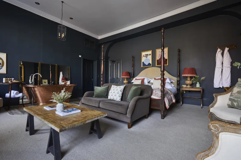 Elegant Kings Head Cirencester suite with four-poster bed, clawfoot tub, gray sofa, and artwork.