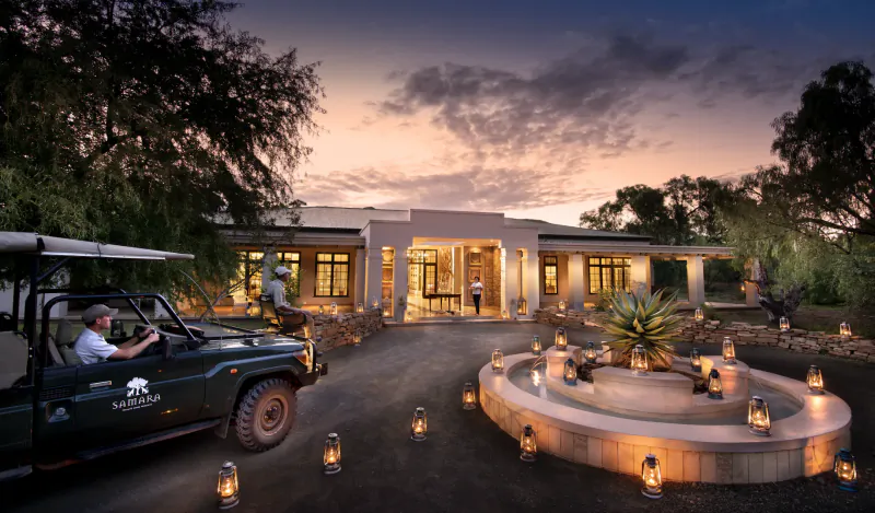Samsara Karoo Safari Villa at dusk, with safari Jeep parked by lantern-lit fountain and entrance courtyard amid trees.