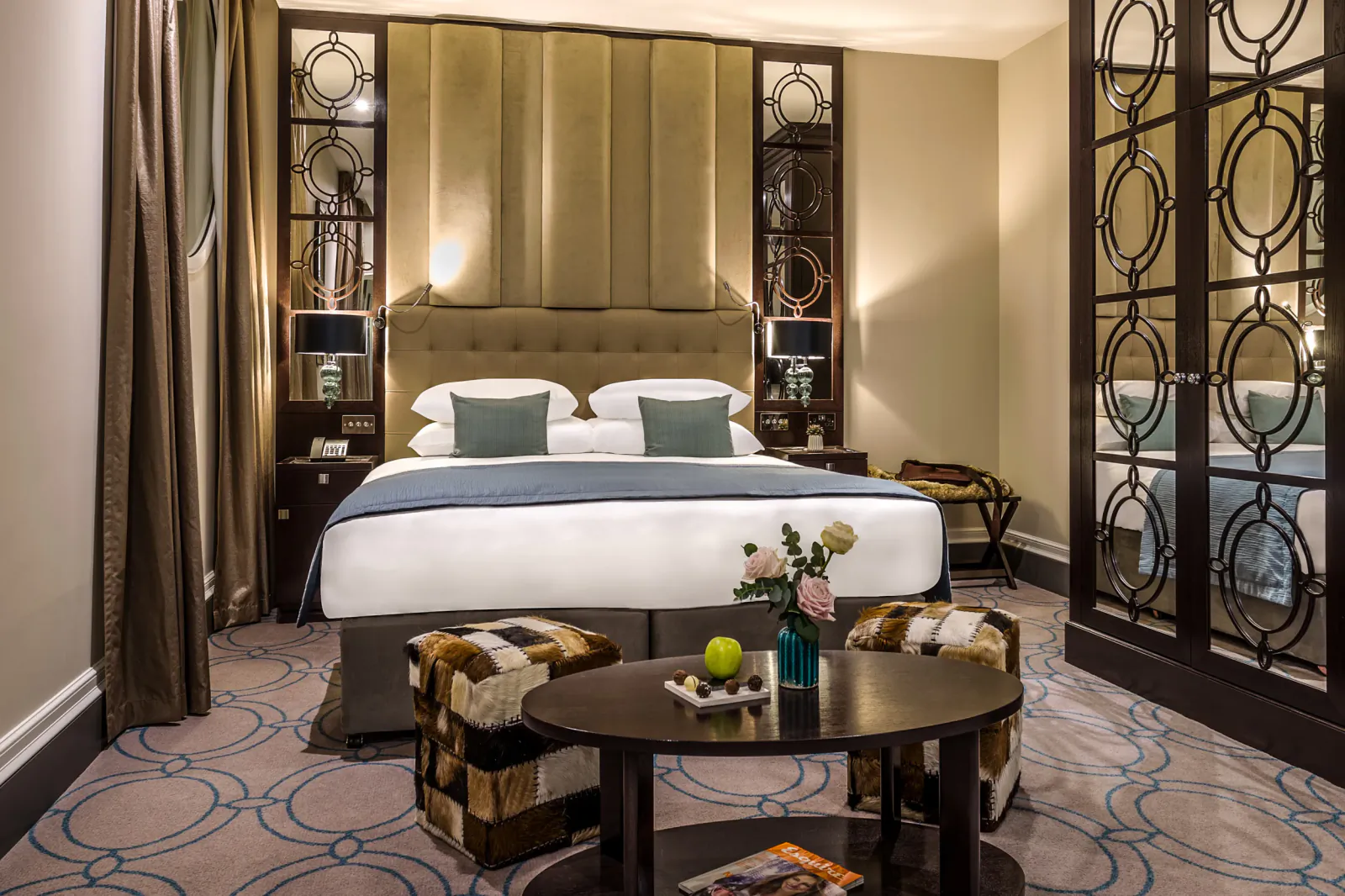 Elegant luxury hotel bedroom with king bed, tufted headboard, ottomans, coffee table with fruit and flowers, ornate screens.