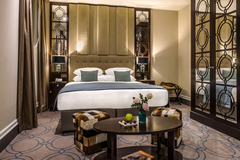 Elegant luxury hotel bedroom with king bed, tufted headboard, ottomans, coffee table with fruit and flowers, ornate screens.