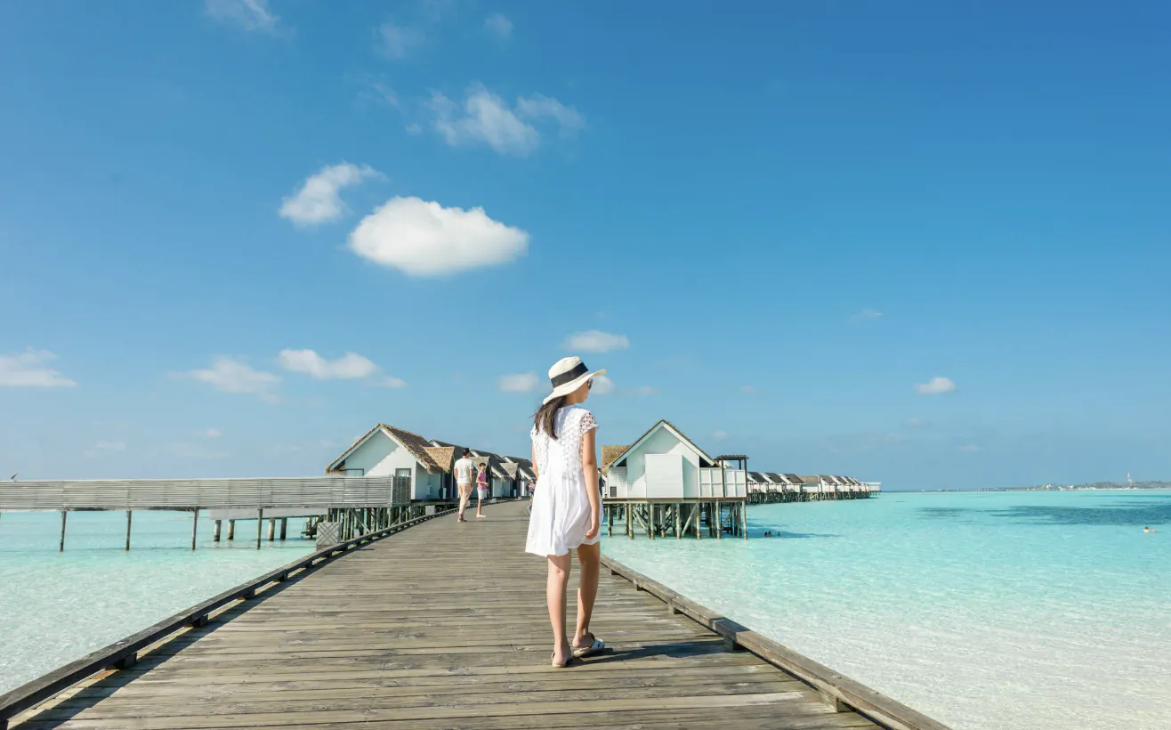Woman in white dress and hat stands on wooden pier facing overwater bungalows and turquoise sea under blue sky.
