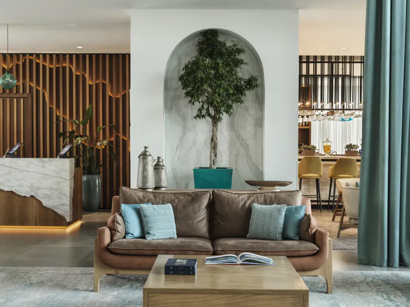 Modern hotel lobby at The Chedi Luštica Bay with wood-paneled walls, marble reception desk, teal sofa, tree in arch, and bar area.