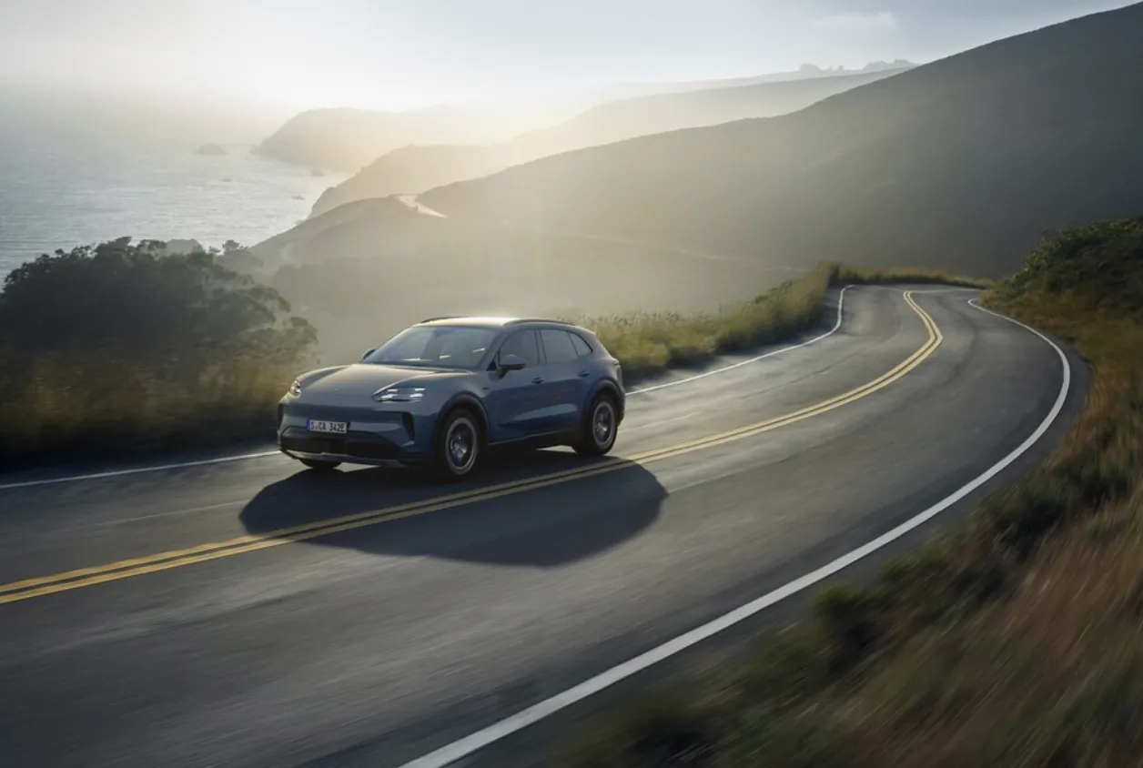 Blue Porsche Cayenne S electric SUV drives on winding coastal road at sunset with ocean and hills.