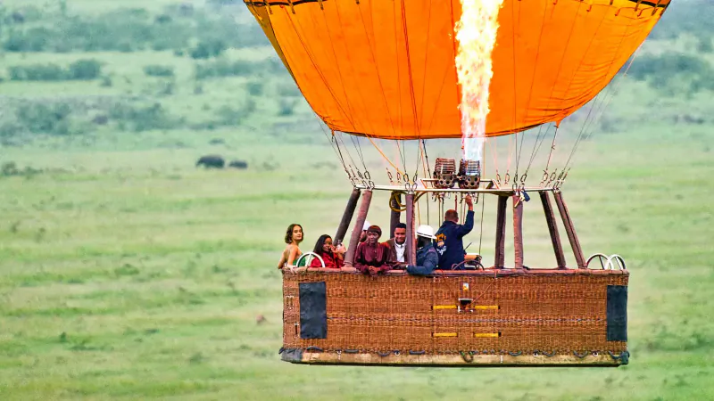 Orange hot air balloon with burner flaming, passengers in basket over Kenyan savanna with distant wildebeest during Great Migration