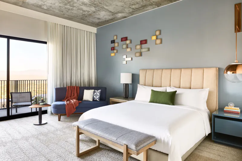 Modern hotel bedroom with king bed, beige headboard, green pillows, loveseat, bench, desert view balcony.