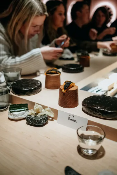Emma Blunt and diners at LUNA Omakase counter with caviar-topped dishes, name tag 'Emma', chopsticks and water glasses on wood surface