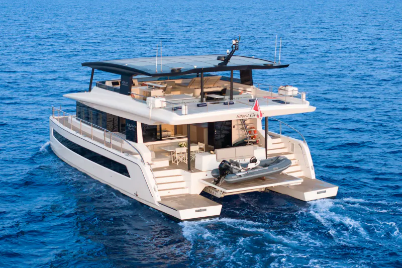 Aft view of luxury white Silent Grace catamaran yacht cruising on blue sea with tender boat and flag.