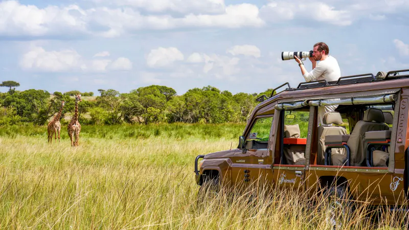 Man photographing giraffes from open-top safari vehicle in Kenyan savanna grassland with acacia trees and clouds.