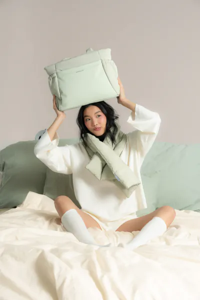 Asian woman sitting on bed, holding up mint green Kapten & Son Cloud Club tote bag, wearing white sweater and socks