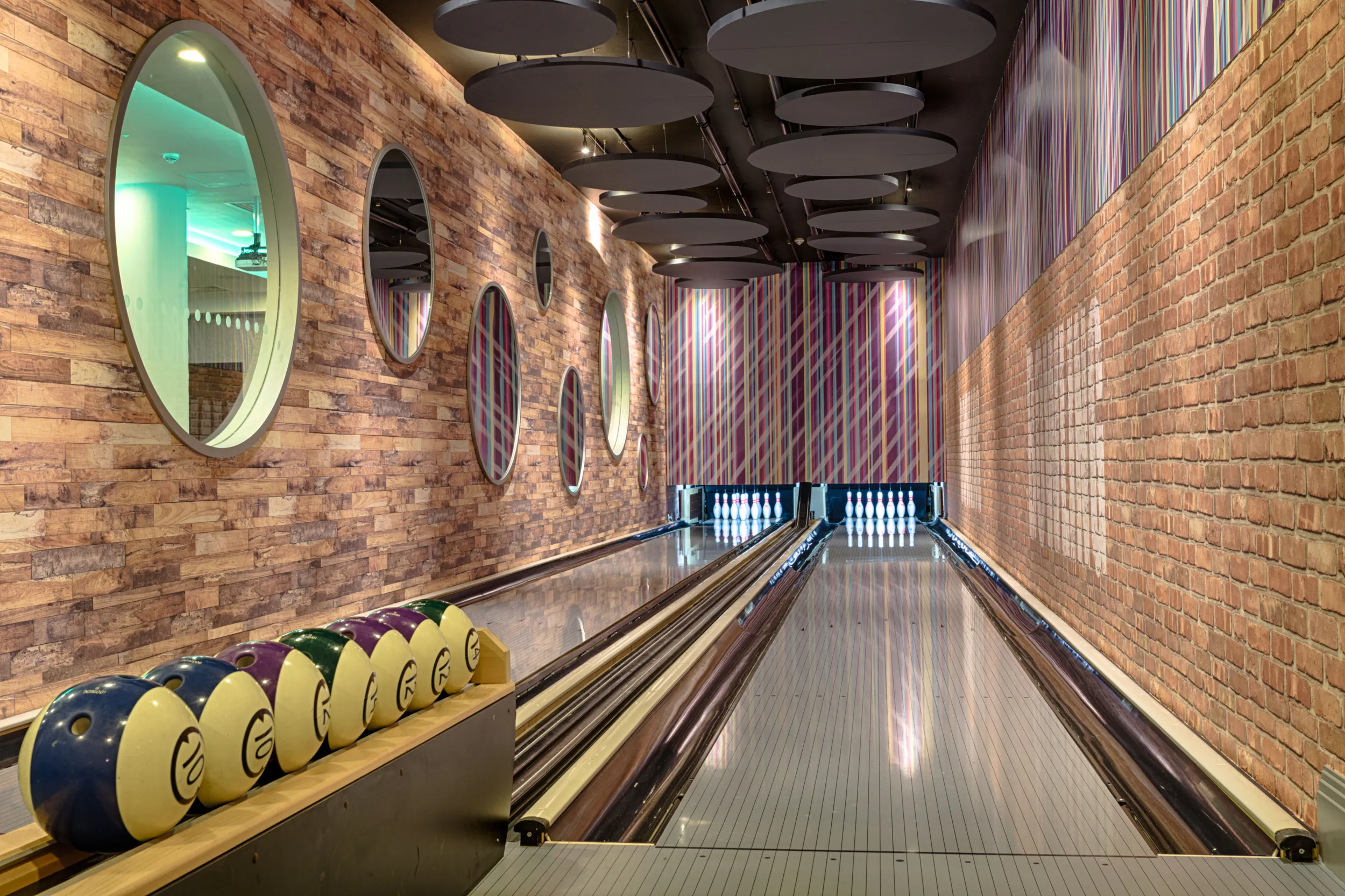 Bowling alley lane with colorful bowling balls ready, exposed brick walls, round mirrors, and striped lighting.