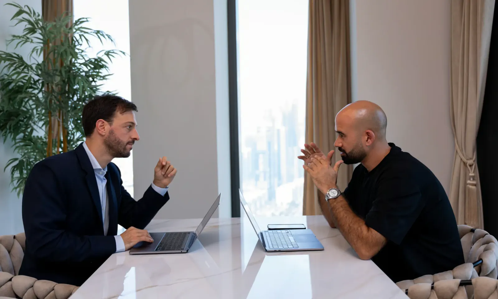 Two businessmen in a modern Dubai office discussing over laptops with city skyline visible through large windows