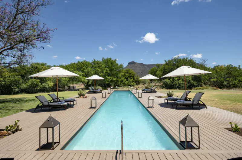 Long turquoise pool at Samara Karoo Safari Villa, surrounded by lounge chairs, lanterns, trees, and mountains under blue sky.