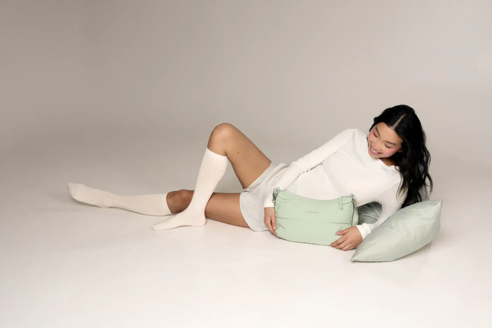 Woman lying on floor in white long-sleeve mini dress and knee-high socks, holding green Kapten & Son Cloud Club pillow