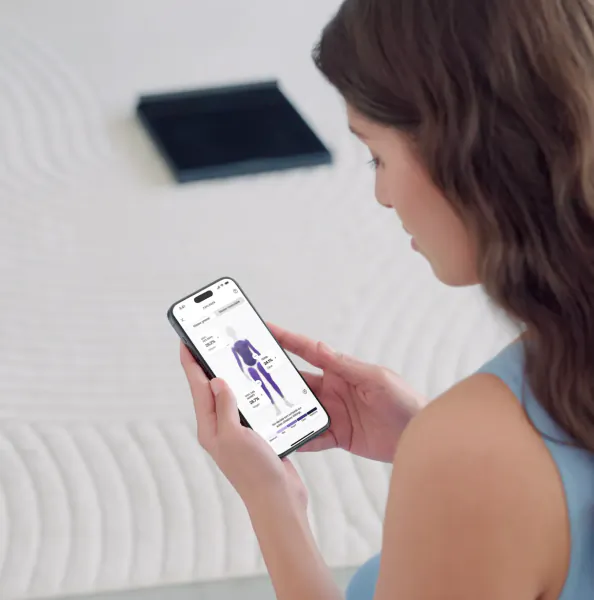 Woman in blue top views 3D human body model on smartphone, standing by bed and black panel.