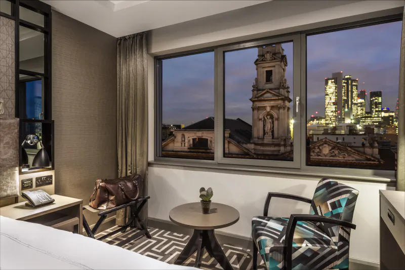 Courthouse Shoreditch hotel room with stylish chairs, plant, and window view of St Leonard's church and London skyline at dusk.