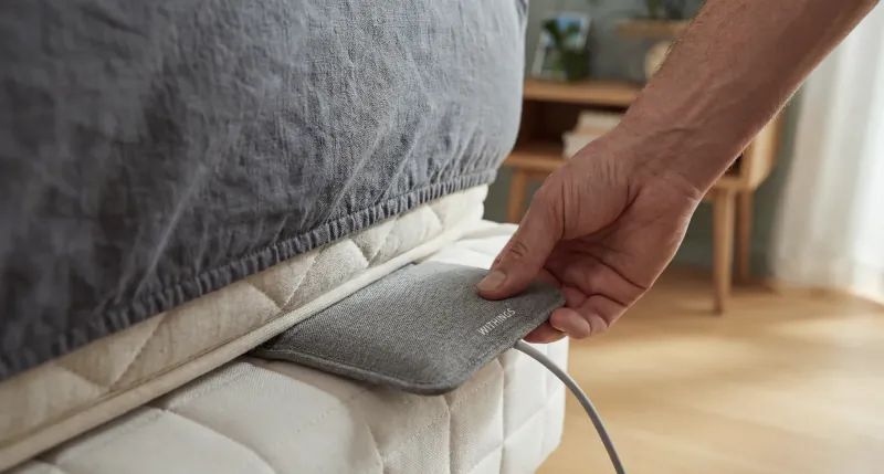 Hand placing grey heated pad under edge of grey bed mattress, connected by cable, in modern bedroom
