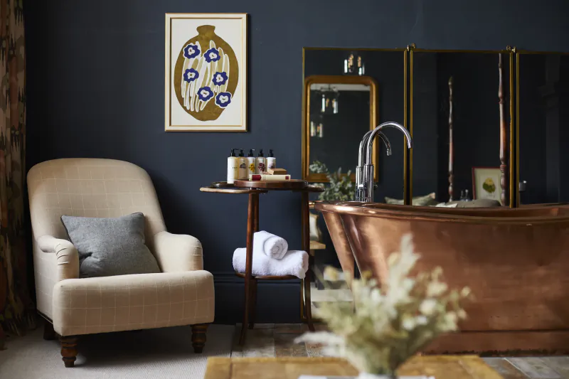 Luxurious bathroom with copper bathtub, beige armchair, towels, bath products, blue flower art, and mirrors at Kings Head Cirencester