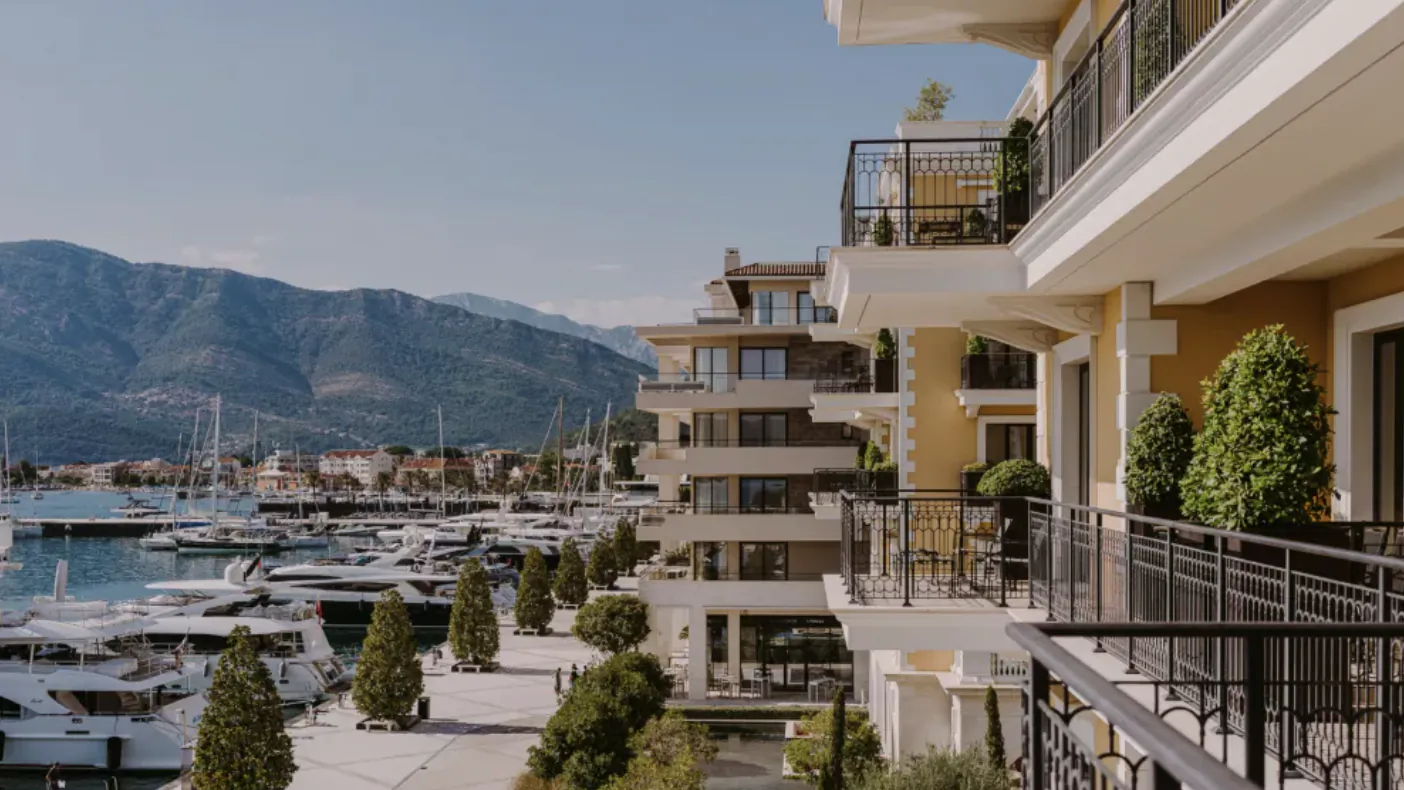 Luxury yellow hotel balconies overlooking marina with yachts, palm trees, and distant mountains under blue sky.