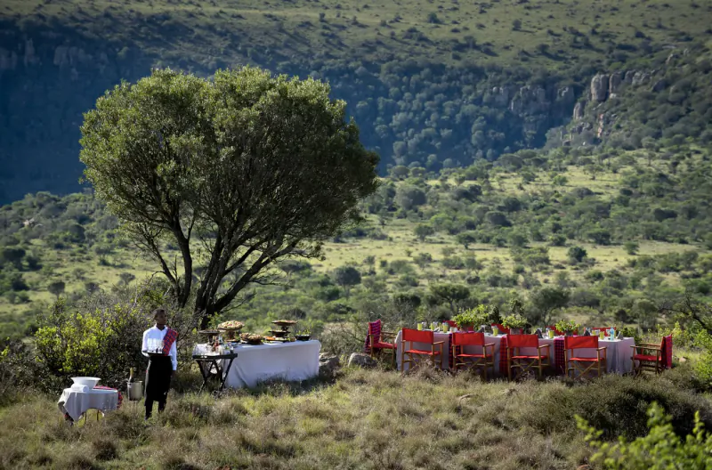 Man in white shirt stands by white-draped table with red chairs under acacia tree in Samara Karoo savanna landscape